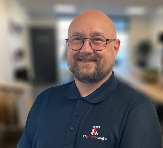 Smilende mand med briller og skæg i mørk polo med logo fra KT Erhvervsbyg A/S