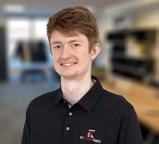 Smilende person i sort poloshirt med logo fra KT Erhvervsbyg A/S foran en sløret kontorbaggrund