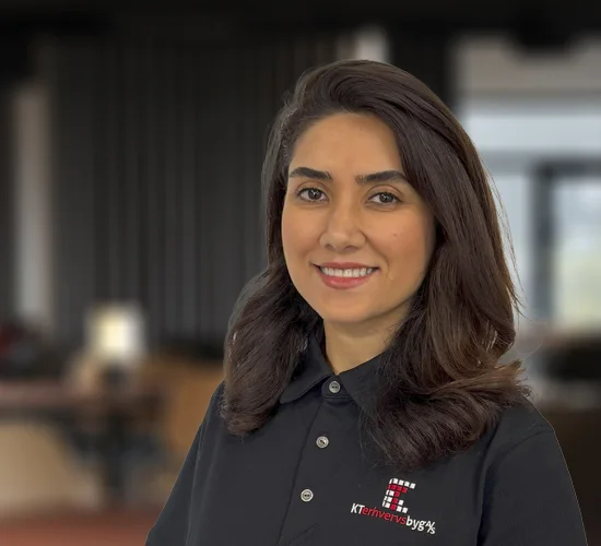 Smilende kvinde med mørkt hår i sort poloshirt med logo fra KT Erhvervsbyg A/S foran en sløret kontorbaggrund