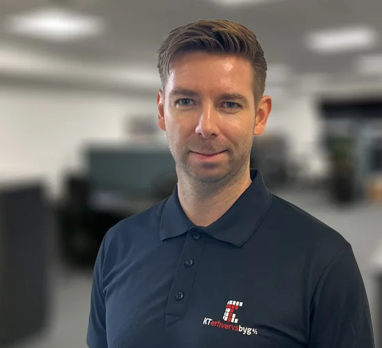 Mand i mørk poloshirt med logo for KT Erhvervsbyg A/S foran en sløret kontorbaggrund