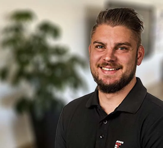 Smilende mand med kort, mørkt hår og skæg i sort poloshirt foran en sløret plante