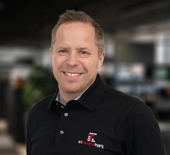 Smilende mand i sort poloshirt med logo for KT Erhvervsbyg A/S foran en sløret baggrund