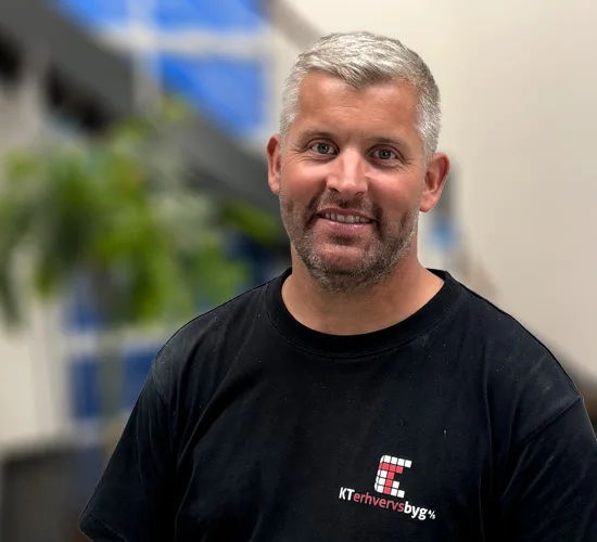 Smilende mand med kort gråt hår og skæg i sort T-shirt med logo fra KT Erhvervsbyg A/S