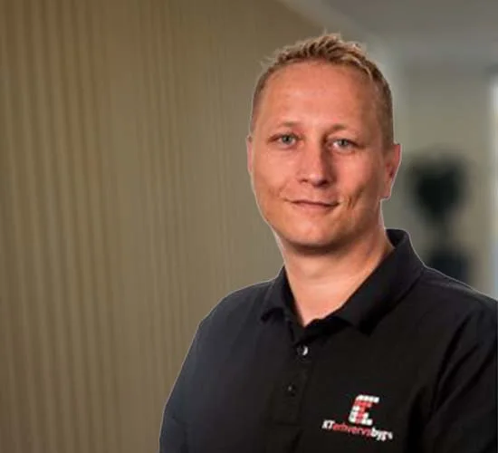 Portræt af en mand med kort, lyst hår og sort poloshirt med firmalogo foran en sløret baggrund