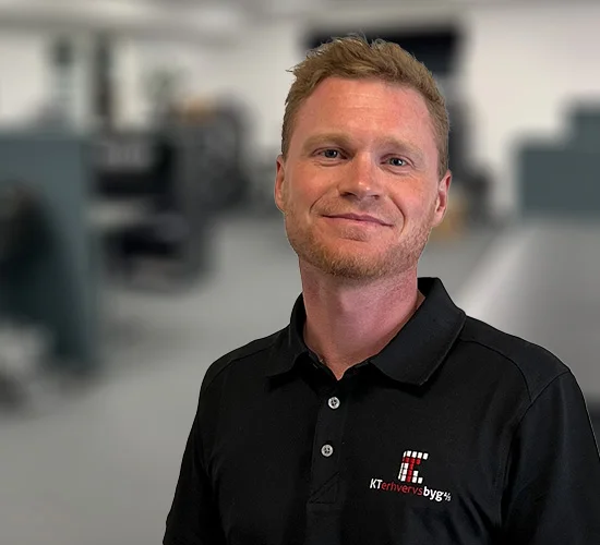 Smilende mand i sort poloshirt med logo for KT Erhvervsbyg A/S foran en sløret baggrund