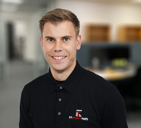 Smilende mand i sort poloshirt med logo for KT Erhvervsbyg A/S foran en sløret baggrund