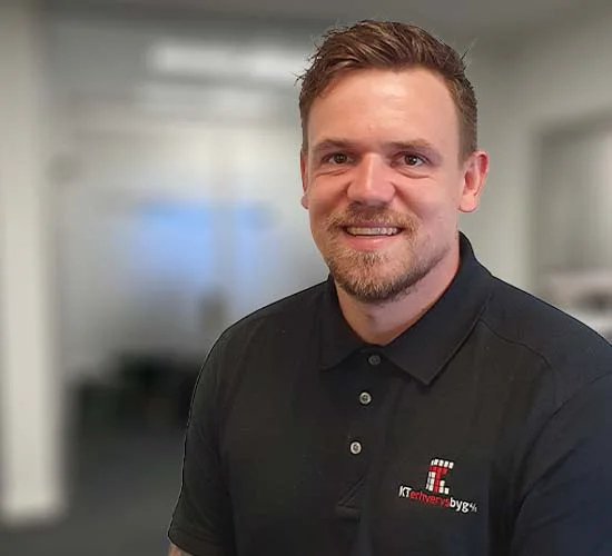 Smilende mand i sort poloshirt med logo fra KT Erhvervsbyg A/S foran en sløret baggrund