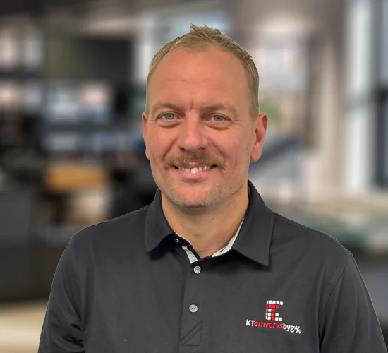 Smilende mand i mørk poloshirt med logo for KT Erhvervsbyg A/S foran en sløret baggrund