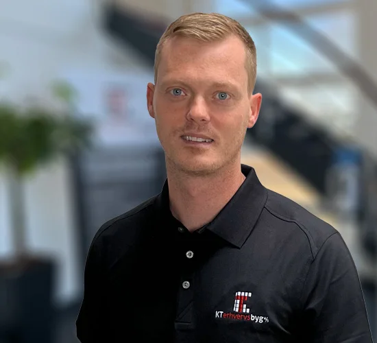 Mand i sort poloshirt med logoet for KT Erhvervsbyg A/S foran en sløret baggrund