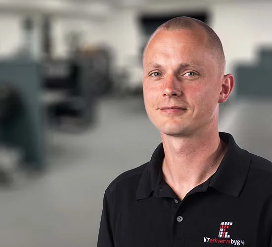 Mand i sort poloshirt med logo fra KT Erhvervsbyg A/S foran en sløret baggrund med maskiner