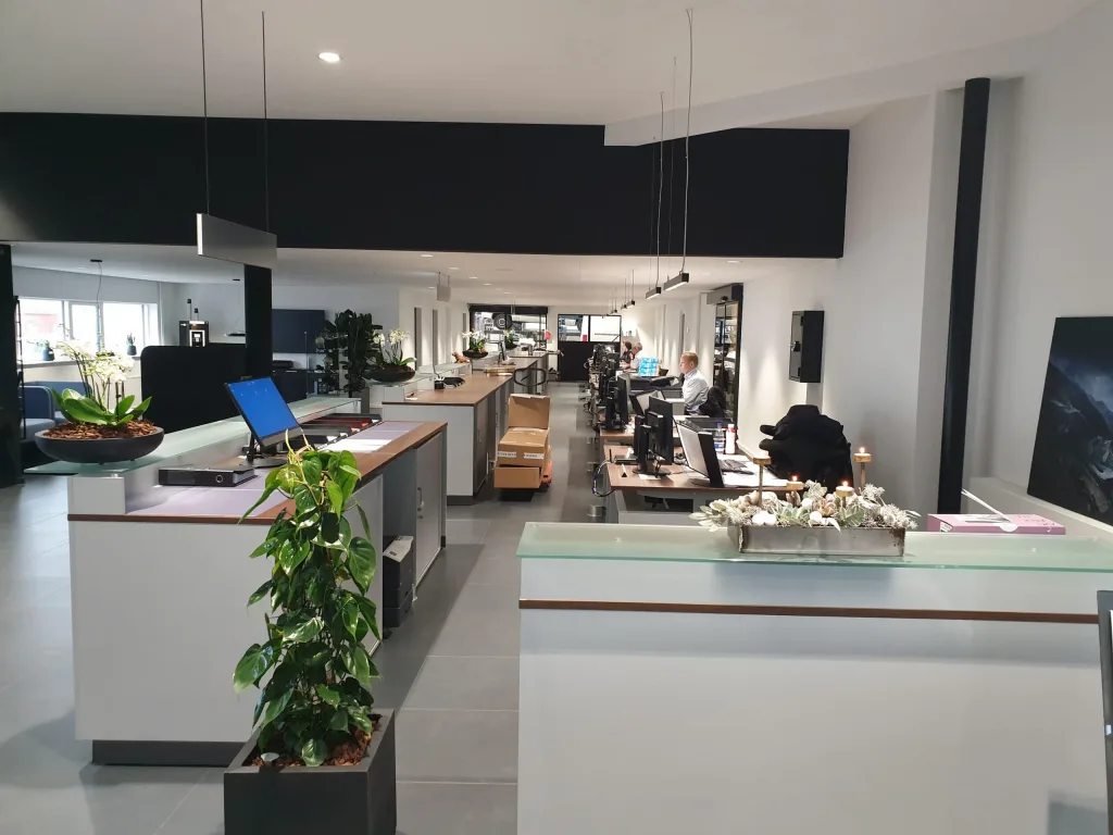Lyst receptions- og kontorområde med skranker, computere, grønne planter og medarbejdere ved skriveborde