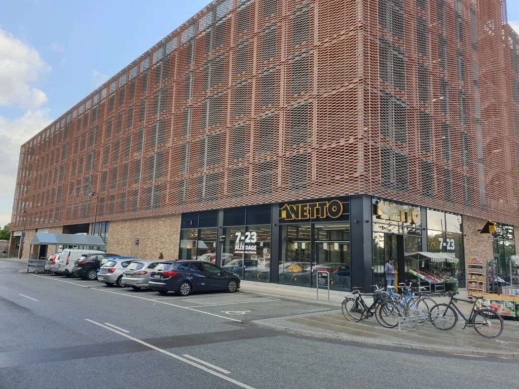 Netto-butik i stueetagen af en stor bygning med mønstret teglfacade, parkerede biler og cykler foran