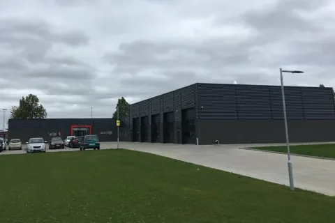 Mørk bygning med flere store garageporte, parkerede biler og en bred indkørsel foran