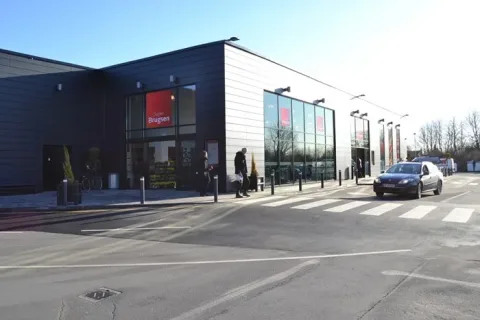 SuperBrugsen med mørk facade og store glaspartier. Foran butikken ses et fodgængerfelt, en bil og nogle gående