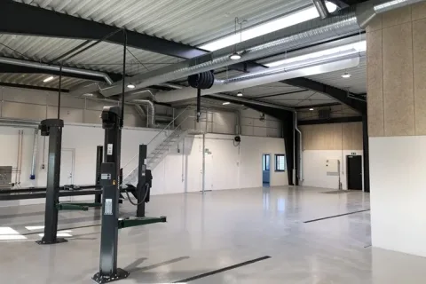 Lyst autoværksted med billifte, ventilationskanaler i loftet og en trappe langs bagvæggen