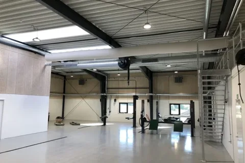 Lyst autoværksted med billifte, synlige ventilationsrør og en ståltrappe langs væggen