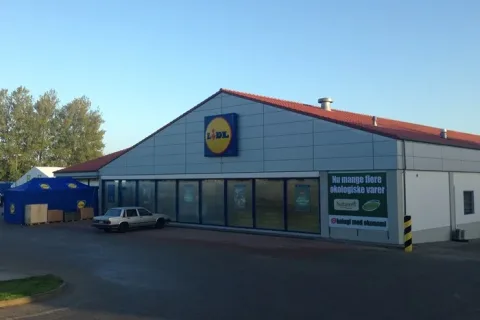 Lidl-butik med grå facade, rødt tag og store vinduer. En bil holder parkeret foran