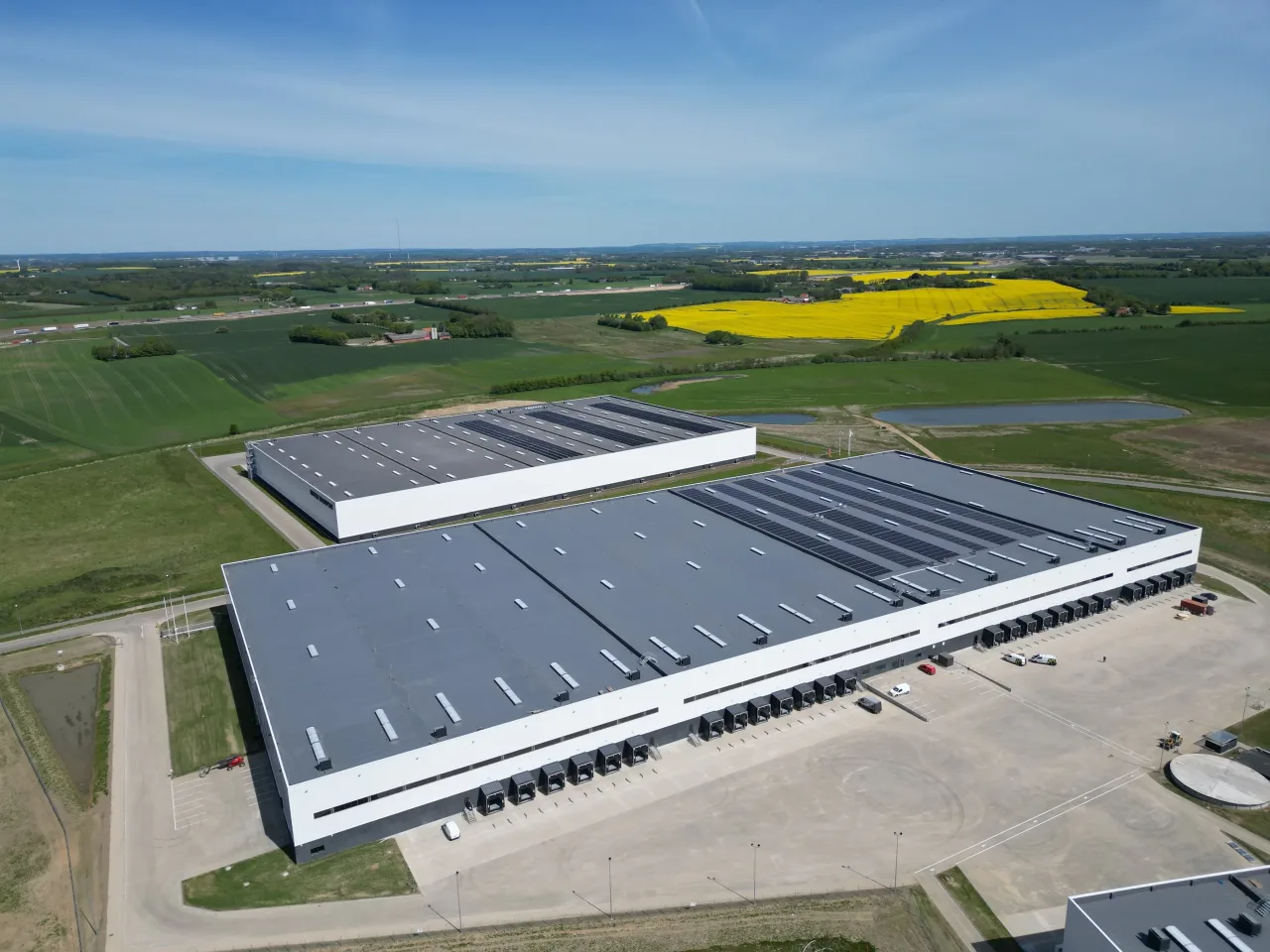 Luftfoto af store lagerbygninger med solceller på tagene, omgivet af grønne marker og gule rapsmarker
