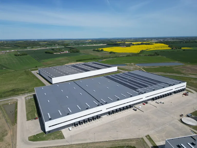 Luftfoto af store lagerbygninger med solceller på tagene, omgivet af grønne marker og gule rapsmarker