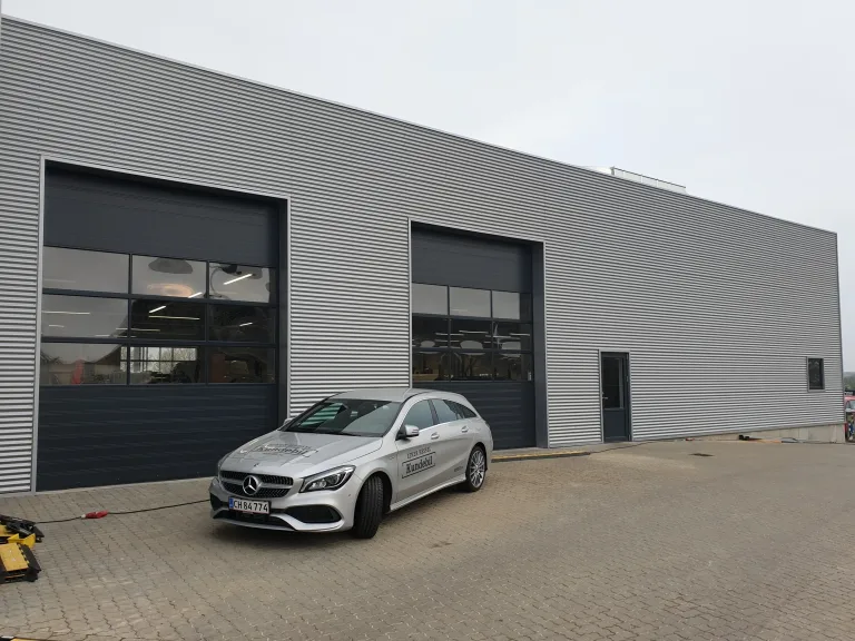 Grå bygning med to store porte med vinduer og en sølvfarvet bil parkeret foran