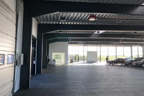 Lys bilhal med højt loft, store glaspartier og flere biler parkeret langs vinduerne