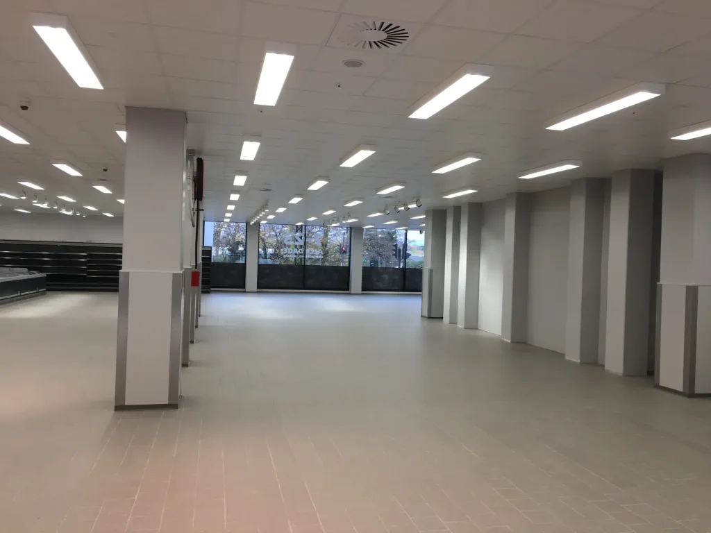 Tomt butikslokale med flisegulv, loftlys, store vinduer og kølemontre langs venstre væg