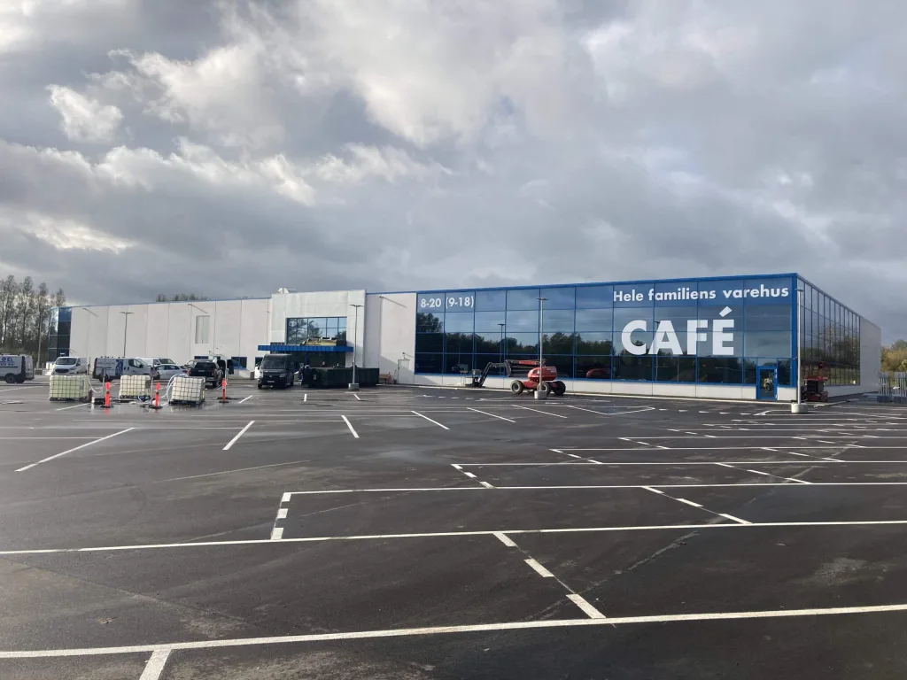 Stort varehus med blå glasfacade og café-skilt foran en næsten tom parkeringsplads med enkelte arbejdskøretøjer