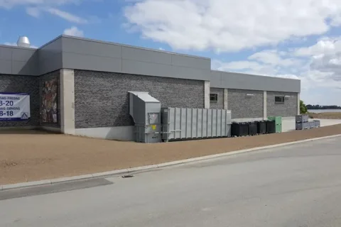 Grå murstensbygning med affaldscontainere og skraldespande langs en asfalteret vej