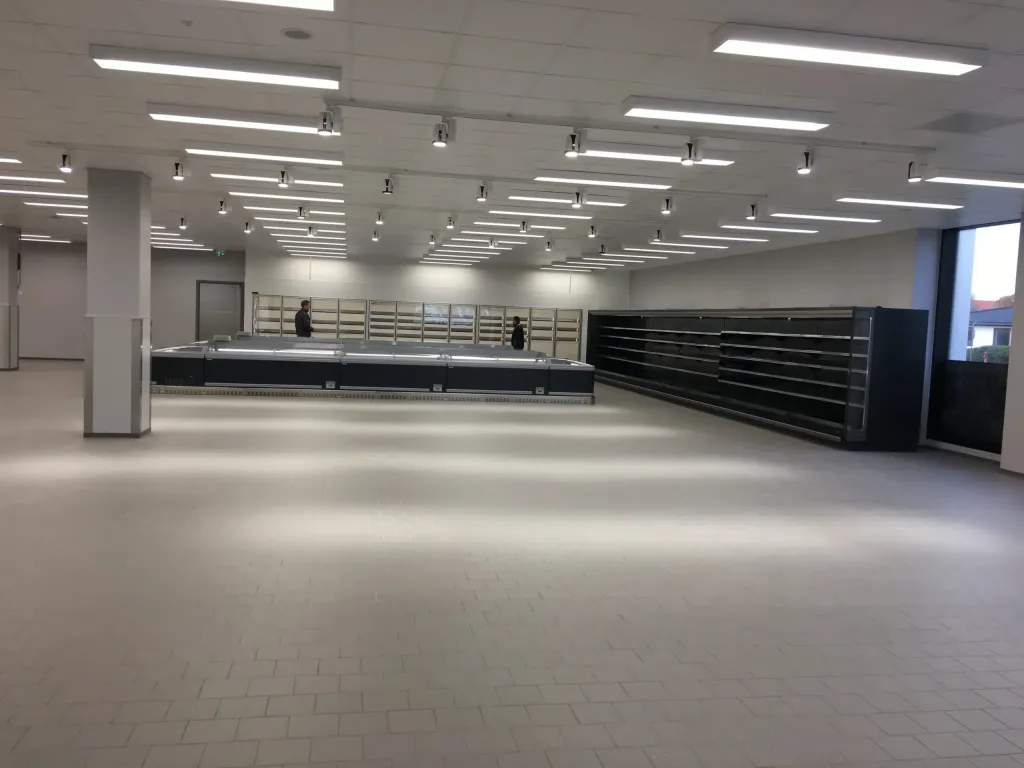 Lyst butikslokale med kølediske, tomme hylder og to personer i baggrunden